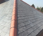 A finished section of a house roof with tidy rows of grey slates and a line of orange ridge tiles along the top.