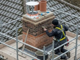 High-definition photo of an active chimney repair on a weathered brick house, showing a technician applying fresh mortar, new bricks installed, scaffolding, and a copper anti-downdraught chimney cowl. Ideal example of professional chimney repair services.