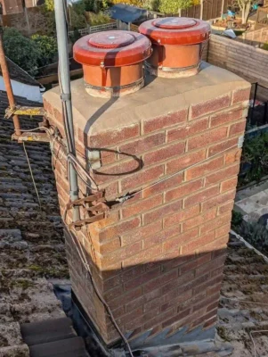 A high-angle shot of a restored brick chimney stack on a residential roof, showing smooth concrete flaunching at the top with two newly installed brown terracotta-coloured anti-down_draught chimney cowls secured by metal straps.