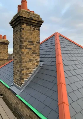 A high-angle shot of a newly installed dark grey slate roof featuring bright orange terracotta hip and ridge tiles, stepped lead flashing around a restored brick chimney stack, and green eaves protection board under an overcast sky.