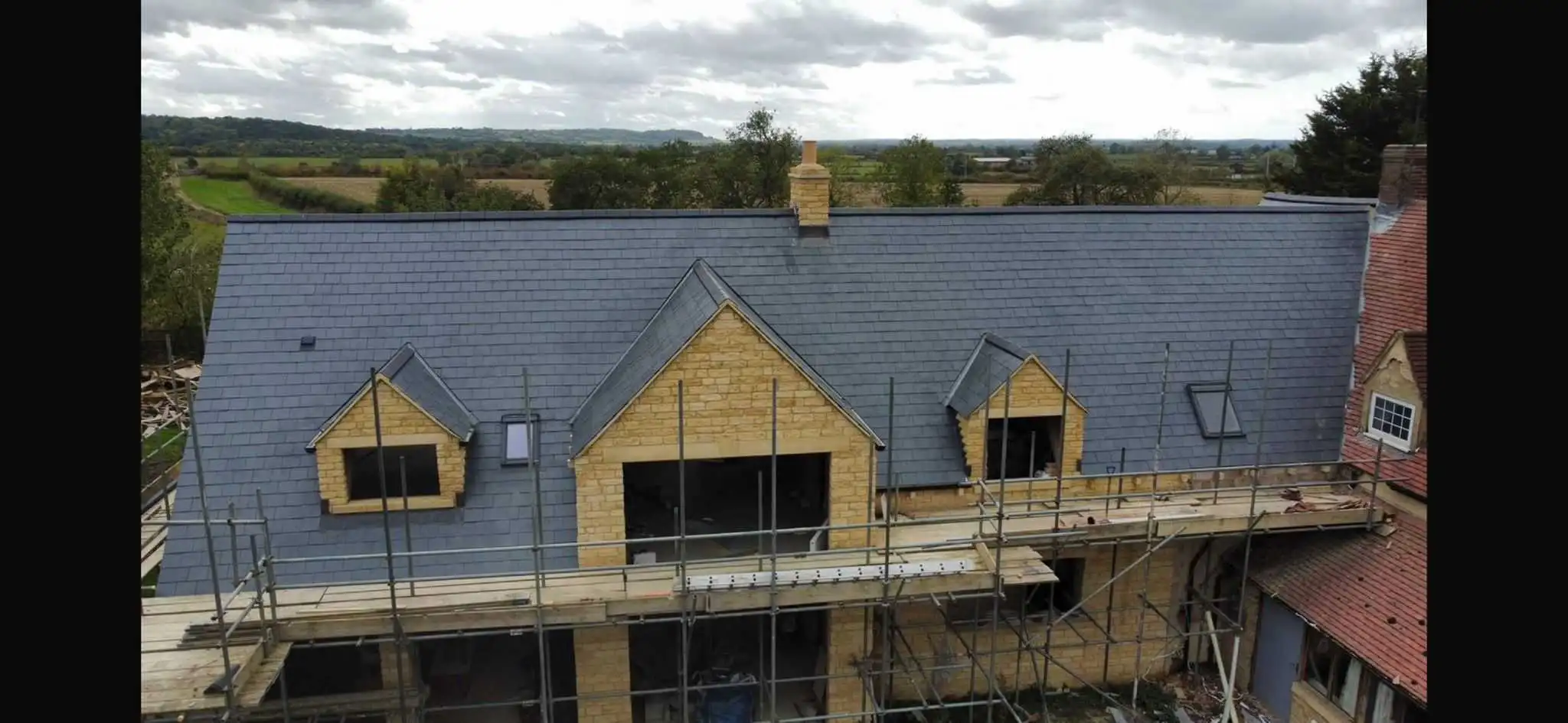 An aerial view of a newly installed dark grey slate roof on a Cotswold stone house under construction, featuring scaffolding, two dormer windows, a central front-facing gable, a stone chimney, and rooflights.