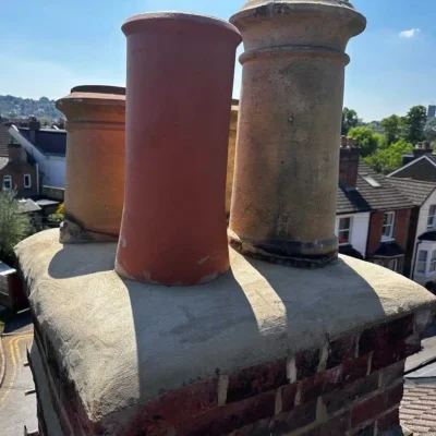 Close-up view of a brick chimney stack undergoing repair, showcasing newly applied smooth concrete flaunching at the base of four traditional terracotta chimney pots against a sunny urban backdrop.