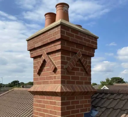 A close-up view of a newly repaired or rebuilt decorative red brick chimney stack on a residential roof with brown concrete tiles. The chimney features ornate corbelled brickwork design elements, clean mortar pointing, terracotta pots, and new stepped lead flashing along the roofline under a partly cloudy sky.