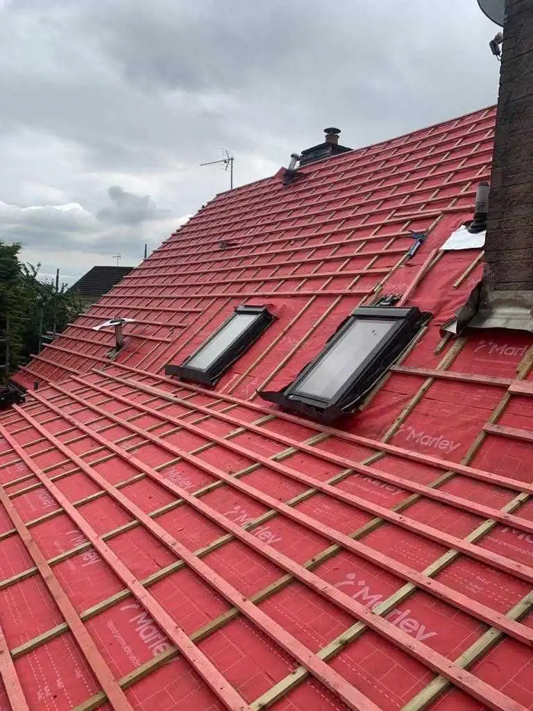 Close-up of a residential roof undergoing repair or re-roofing, showing red Marley breathable underlay membrane, freshly installed timber tiling battens, and two integrated Velux skylight windows.