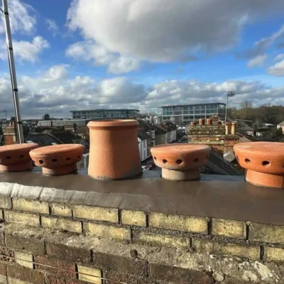 A close-up of a repaired brick chimney stack on a terraced house, showing fresh grey mortar flaunching with an array of terracotta chimney pots, including three round pepperpot ventilation cowls, overlooking a UK town under a blue sky.
