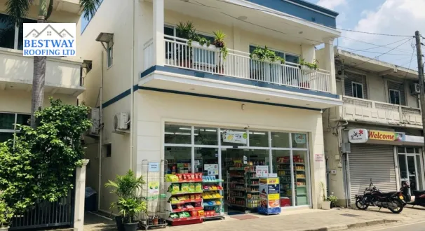A two-story cream-colored commercial building featuring a convenience store on the ground floor, a white-railed balcony on the upper level, and a flat roofline with deep blue architectural trim and visible drainage downpipes.
